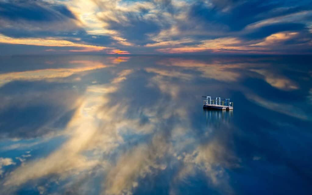 A small boat floats on a calm, reflective body of water under a dramatic sky with wispy clouds and sunset hues.