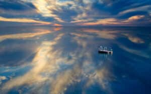 A small boat floats on a calm, reflective body of water under a dramatic sky with wispy clouds and sunset hues.